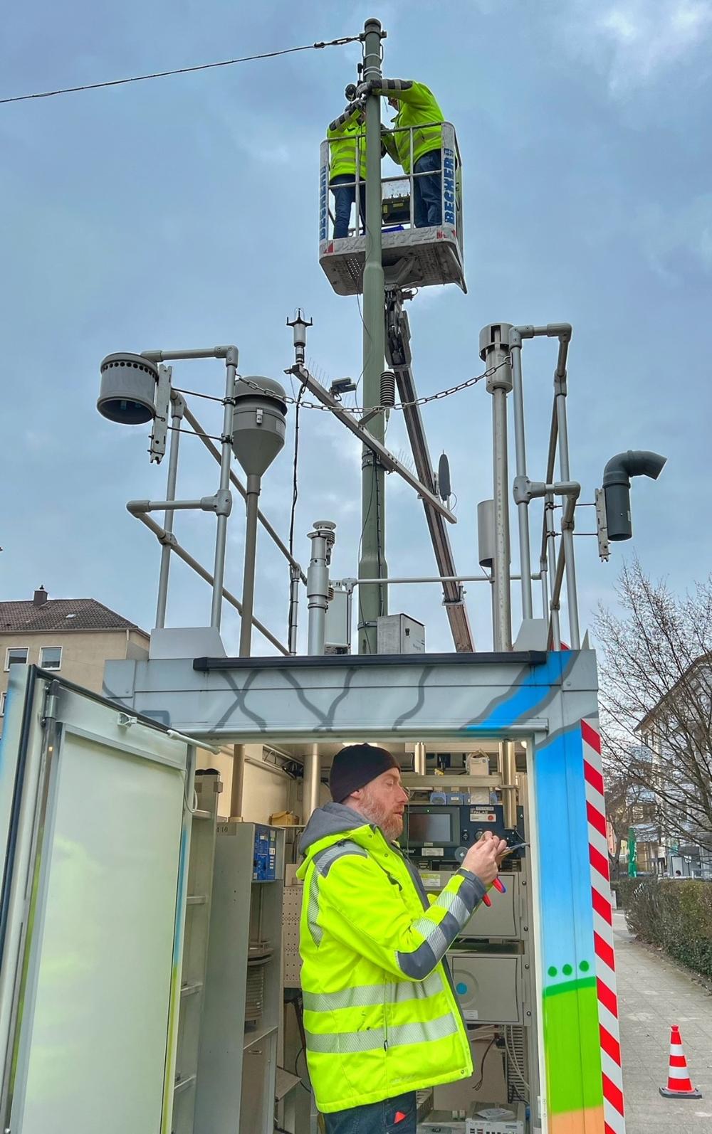 Ein Mann steht in der Luftmessstation unten und installiert die Datenübertragung, ob auf einem Hubsteiger installieren zwei Mitarbeiter die Sensoren