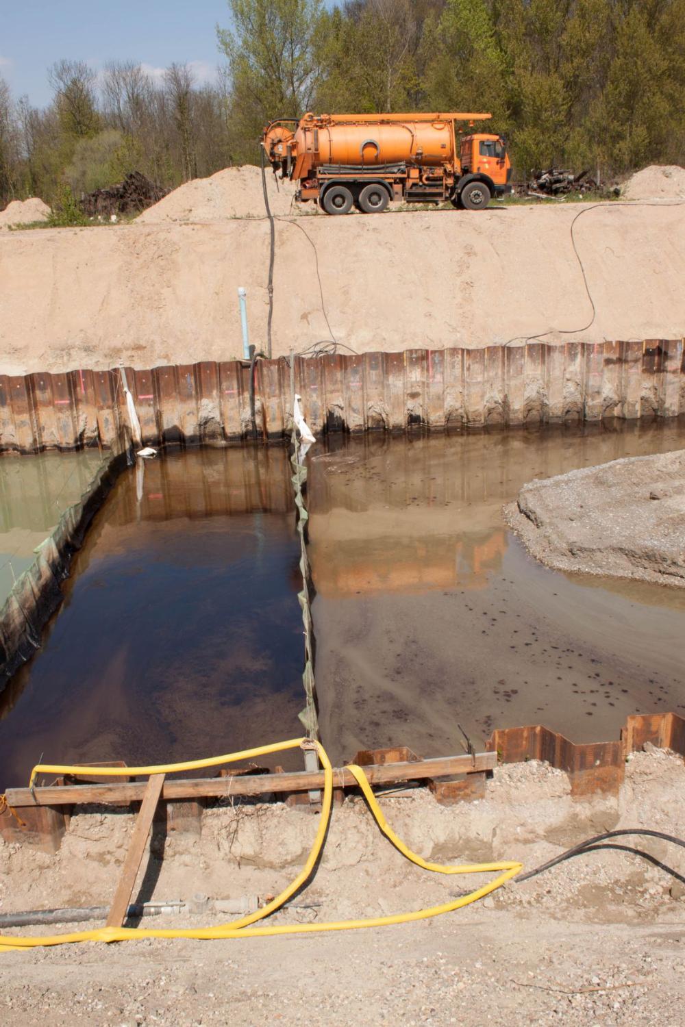 Umspundeter Bereich von mit Mineralölkohlenwasserstoffen belastetem Grundwasser. Das Wasser schimmert schwarz-ölig. Im Hintergrund ist ein Pumpfahrzeug auf einer Anhöhe. 