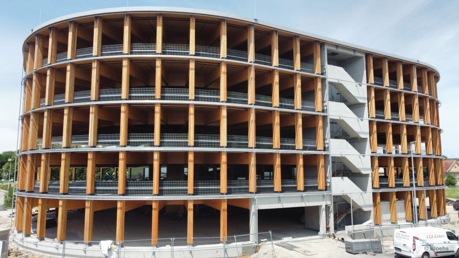 Das Holzparkhaus hat eine ovale Grundform und 5 Stockwerke. Jedes Stockwerk hat eine Höhe von ca. 3 Meter. Man blickt von links vorne im Winkel von ca. 45°auf die Rundung des Ovalgebäudes. Das Gebäude ist noch im Rohbau, aber fast fertig. Jedes Stockwerk ist mit Holzbalken abgestützt, die sich im Abstand von ca. 5 Meter befinden. Im mittleren Bereich ist die Einfahrt - und rechts daneben das Treppenhaus in Betonbauweise, beides noch im Rohbau zu sehen. In der rechten unteren Ecke steht ein weißer Kastenwagen mit der Firmenaufschift 