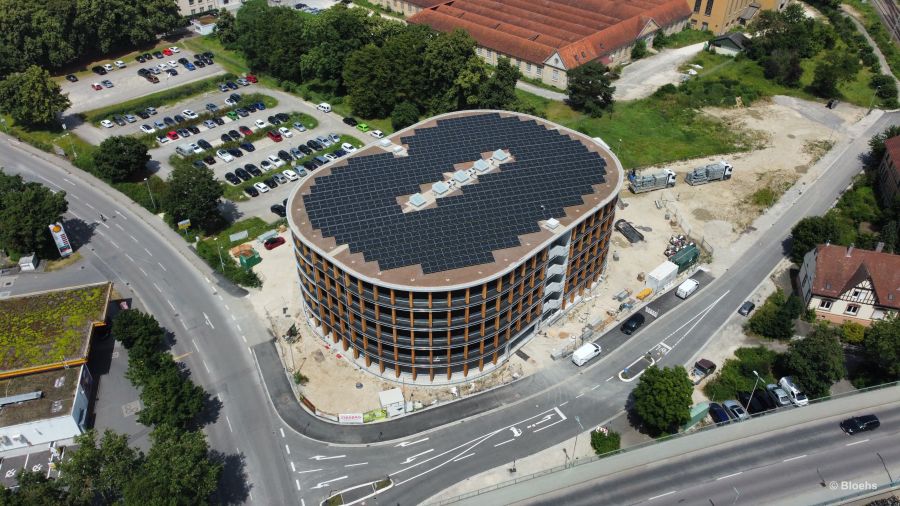 Man blickt aus ca. 30 Meter Höhe im diagonalen Winkel (ca. 45°) auf das Gebäude. In der Mitte des Dachs sind parallel zur Längsseite 5 Dachfenster hintereinander angebracht. Die Dachfläche ist fast vollständig mit Photovoltaik-Modulen bestückt. Parallel zur hinteren Längsseite befindet sich ein Parkplatz. Parallel zur vorderen Längsseite führt die Zufahrtsstraße zum Parkhaus vorbei. An der linken Schmalseite führt eine Straße vorbei.