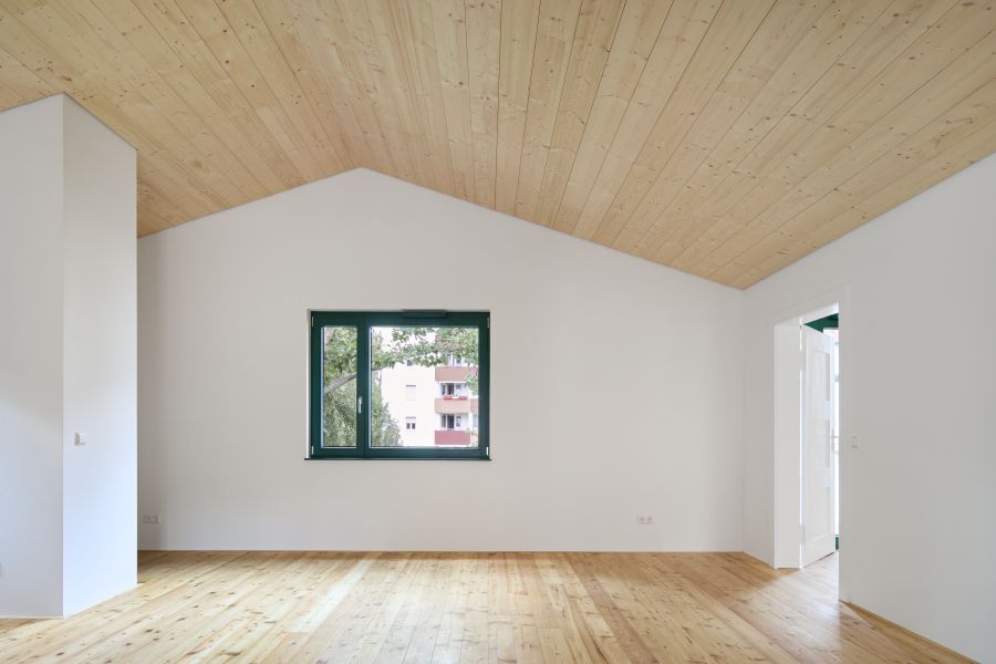  Wohnraum mit Parkettboden, Blick auf die Außenwand mit eingelassenem Fenster. Die Giebeldecke ist mit Holzpanälen verkleidet.