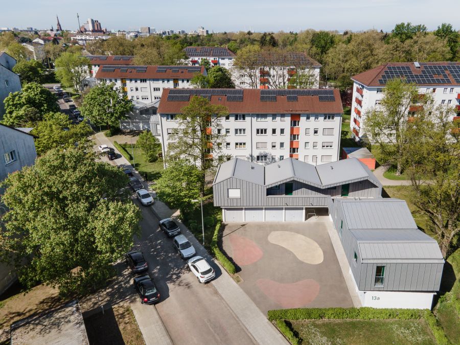  Luftansicht aus ca. 50 Meter Höhe schräg auf die Garagenaufstockung. Links daneben die Straße mit parkenden Autos. Im Hintergrund mehrere vier- bis fünfstöckige Wohnblocks mit Giebeldächern, die mit Photovoltaikmodulen bestückt sind.