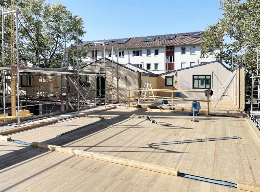  Baustelle Bodenplatte aus Holz, seitlich ein Gerüst, im Hintergrund steht quer zur Baustelle ein Mehrgeshossiges Wohnhaus, auf dessem Dach Photovoltaik-Module installiert sind.