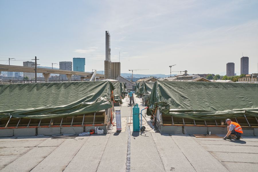 Blick auf das Dach während der Sanierung. Aufgesetzte Dachgauben mit grünen Planen abgedeckt. Im Vordergrund der Bildmitte steht eine türkisfarbene Gasflasche auf einem Gasflaschenwagen, links daneben stehend eine Dachpappenrolle. Rechts im Vordergrund ein Bauarbeiter in orangefarbener Warnweste, in der Bildmitte ein Arbeiter stehend mit blauem T-Shirt. Im Hintergrund links der Bildmitte vier Hochhäuser sowie ein die Hochhäuser überragender Turm. Am linken Bildrand im Hintergrund zwei etwa gleichhohe quadratische Hochhäuser. Vom linken Bildrand bis in den Hintergrund der Bildmitte erstreckt sich eine Hochstraße in einer langezogenen Linkskurve. Blick auf das Dach während der Sanierung. Aufgesetzte Dachgauben mit grünen Planen abgedeckt. Im Vordergrund der Bildmitte steht eine türkisfarbene Gasflasche auf einem Gasflaschenwagen, links daneben stehend eine Dachpappenrolle. Rechts im Vordergrund ein Bauarbeiter in orangefarbener Warnweste, in der Bildmitte ein Arbeiter stehend mit blauem T-Shirt. Im Hintergrund links der Bildmitte vier Hochhäuser sowie ein die Hochhäuser überragender Turm. Am linken Bildrand im Hintergrund zwei etwa gleichhohe quadratische Hochhäuser. Vom linken Bildrand bis in den Hintergrund der Bildmitte erstreckt sich eine Hochstraße in einer langezogenen Linkskurve.