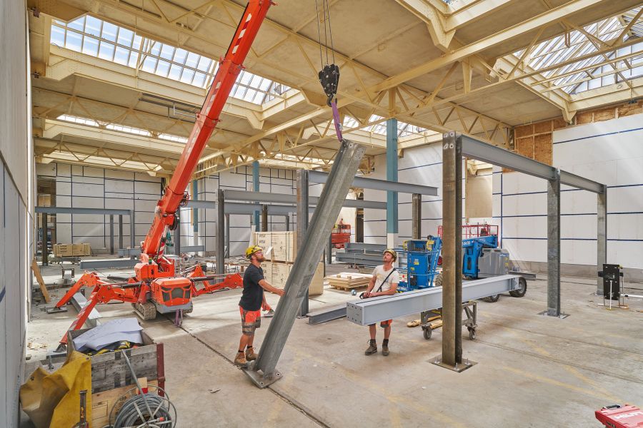 Blick in die Halle, in der von Zwei Arbeitern die Stahlträger montiert werden. Als Montagehilfe dient ein mobiler, orangefarbener Lastenkran. Das Dach ist eine Holzkonstruktion mit Oberlichtern. Blick in die Halle, in der von Zwei Arbeitern die Stahlträger montiert werden. Als Montagehilfe dient ein mobiler, orangefarbener Lastenkran. Das Dach ist eine Holzkonstruktion mit Oberlichtern.