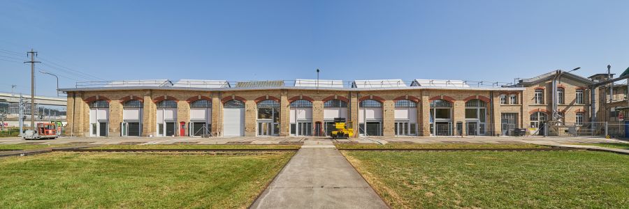 Blick auf die Bauwerksfassade. Die Fassade besteht aus hellen Backsteinen. Im Flachbau sind 12 Fensterelement angebracht, die vom Boden bis unter das Dach reichen. Die Oberseite der Fensterelemente sind Rundbögen. Auf dem Dach sieht man die in Querrichtung aufgesetzten giebelförmigen Oberlichter. Rechts neben dem Flachbau ist ein etwas höheres Gebäude mit Giebeldach zu sehen. Blick auf die Bauwerksfassade. Die Fassade besteht aus hellen Backsteinen. Im Flachbau sind 12 Fensterelement angebracht, die vom Boden bis unter das Dach reichen. Die Oberseite der Fensterelemente sind Rundbögen. Auf dem Dach sieht man die in Querrichtung aufgesetzten giebelförmigen Oberlichter. Rechts neben dem Flachbau ist ein etwas höheres Gebäude mit Giebeldach zu sehen.