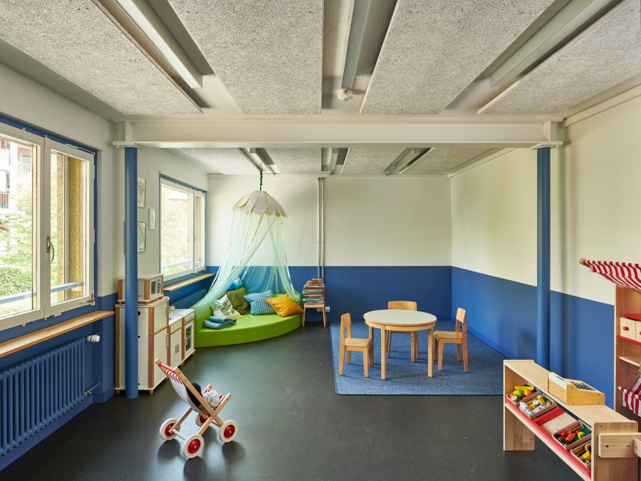 Blick in das Kindergartenzimmer. Der untere Bereich der Wände ist blau, links die Fensterseite. An der Decke sieht man den in Querrichtung angebrachten Stahlträger und die in Längsrichtung ausgerichteten Deckenpanele