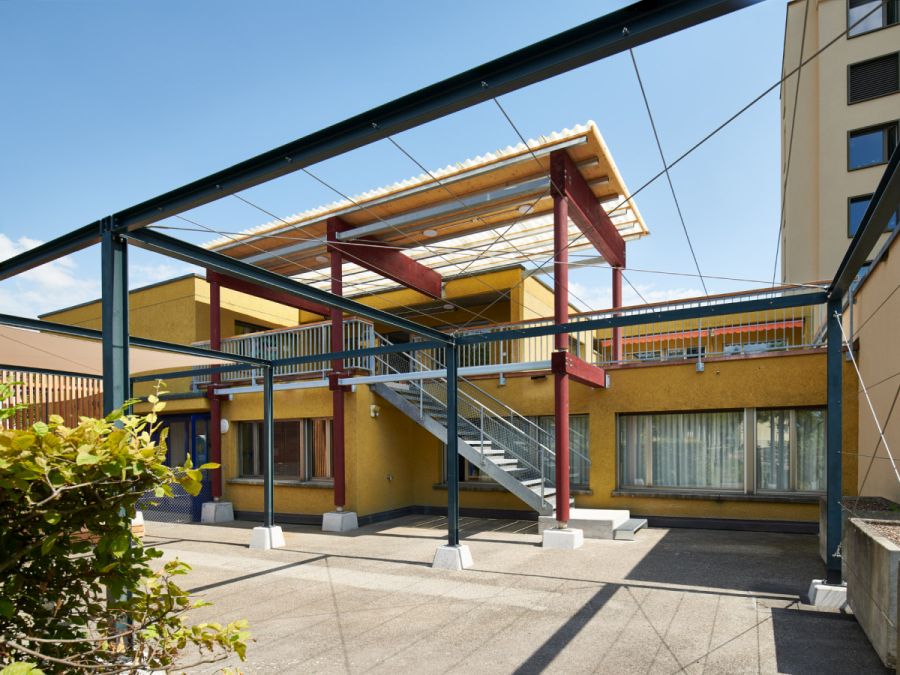  Außenansicht des Kindergartens. Zum Obergeschoss führt eine Stahltreppe. Der obere Terrassenbereich ist mit einer Pergola in Holzkonstruktion abgedeckt.