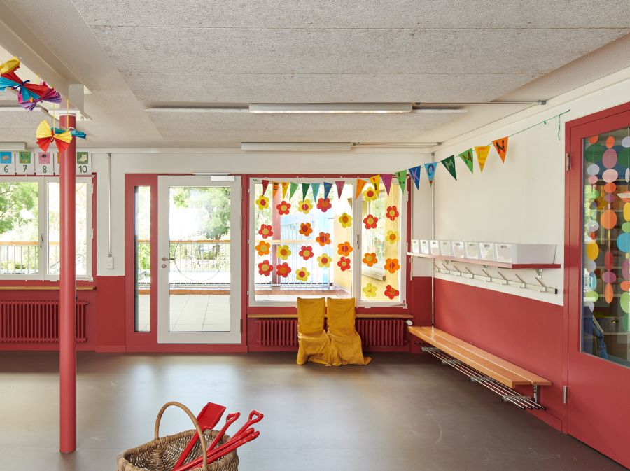 Im lichtdurchfluteten Eingangsbereich fallen die im unteren Drittel rot gestrichenen Wände auf. Rechts an der Wand ist eine Sitzbank montiert. Oberhalb der Sitzbank ist die Garderobe, sowie ein Regal, auf dem acht weiße Kunststoffschalen abgestellt sind. Das Fenster ist mit orange-/gelben Papierblumen verziert.