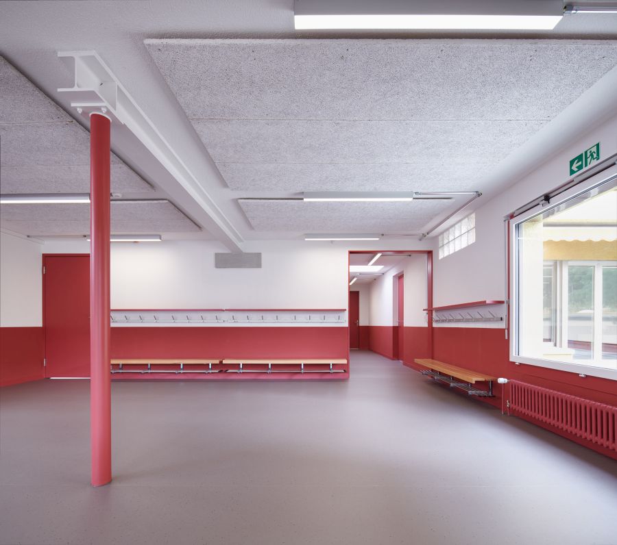  Blick in den Eingangsbereich. Die Wände im unteren Bereich sind rot gestrichen. An der Decke ist im Längsbereich ein Stahlträger zu sehen. Rechts das Fenster zum Außenbereich. Unterhalb des Fensters der ebenfalls in rot gestrichene Heizkörper. An den Wänden sind Sitzbänke montiert, darüber befinden sich die Garderoben.