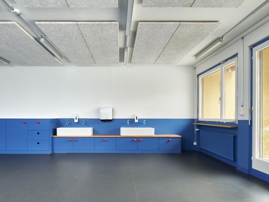 Blick in den Waschraum. Der untere Bereich der Wände ist blau gestrichen. Der obere Bereich der Wände, sowie die Decke ist weiß. Rechts vorne die Terrassentür, dahinter das Fenster, unter dem eine Holzfensterbank und darunter ein in ebenfalls blau gestrichener Heizkörper. Die Schränke sind ebenfalls in Blau gestrichen. Auf den Waschtischen stehen zwei weiße, rechteckige Waschbecken.