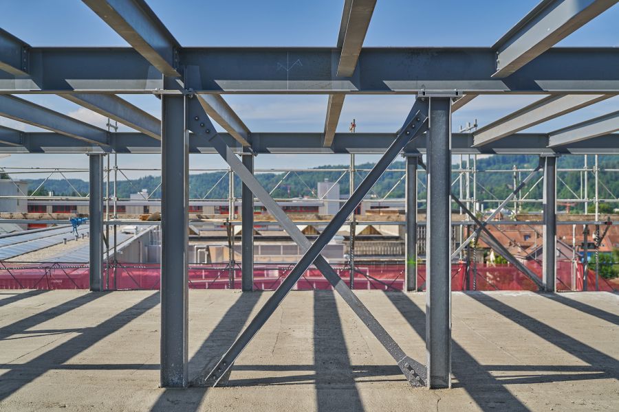 Das Bild zeigt eine Baustelle, genauer gesagt den Rohbau eines Gebäudes aus einer Stahlkonstruktion. Im Vordergrund sind mehrere senkrechte Stahlträger und horizontale Querträger zu sehen, die ein stabiles Gerüst bilden. Besonders auffällig ist eine diagonale Kreuzverstrebung (in X-Form), die zur Aussteifung der Struktur dient und für zusätzliche Stabilität sorgt. Der Boden besteht aus einer glatten Betondecke, und rundherum ist ein Baugerüst mit Sicherheitsnetzen und Absperrungen (in Rot) zu erkennen. Im Hintergrund sieht man Dächer anderer Gebäude, Bäume und Hügel, was auf eine Lage am Rande einer Stadt oder in einer industriellen Umgebung mit landschaftlichem Hintergrund hinweist. Der Himmel ist klar und blau, was auf gutes Wetter während der Bauarbeiten schließen lässt.