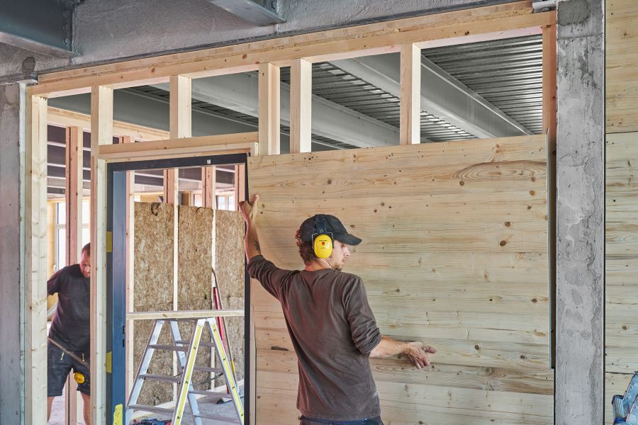 Das Bild zeigt eine Innenaufnahme einer Baustelle, auf der zwei Handwerker beim Errichten einer Innenwand arbeiten. Im Vordergrund steht eine Person mit gelben Gehörschutzkopfhörern und einer dunklen Kappe. Sie hält eine große Holzplatte, die gerade an eine Wandkonstruktion aus Holzrahmen montiert wird. Diese Holzrahmenkonstruktion dient als Unterbau für die Wandverkleidung. Im Hintergrund ist ein weiterer Arbeiter zu sehen, der ebenfalls mit Holz arbeitet. Neben ihm steht eine Leiter, und man erkennt Dämmmaterial sowie Werkzeuge, die für den Innenausbau verwendet werden. Die Umgebung besteht aus einer Mischung aus Beton, Stahl und Holz – typische Materialien beim Umbau oder der Renovierung eines Industriegebäudes. Das Licht im Raum ist hell und natürlich, vermutlich durch große Fenster oder eine offene Gebäudestruktur.