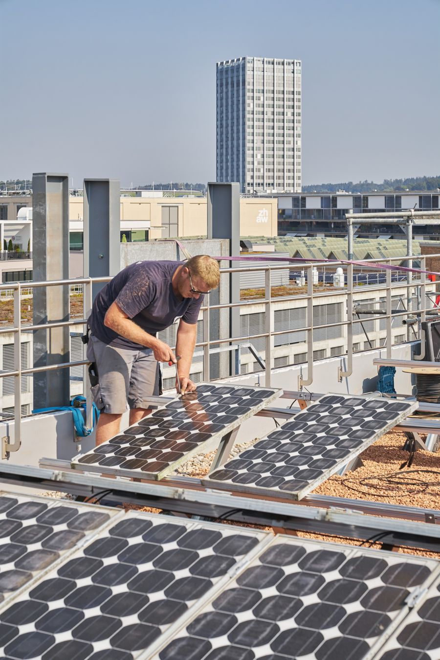 Das Bild zeigt einen Arbeiter, der auf einem Flachdach Solarmodule installiert. Im Vordergrund sind mehrere Solarpaneele zu sehen, die schräg auf Metallgestellen montiert sind. Der Mann trägt ein dunkles T-Shirt, graue Shorts und eine Sonnenbrille. Er arbeitet konzentriert mit einem Werkzeug, vermutlich einem Schraubenzieher, an einem der Module. Der Boden ist mit Kies bedeckt, wie es bei Flachdächern häufig der Fall ist. Im Hintergrund sind moderne Gebäude und ein hoher Turm zu erkennen. Eines der Gebäude trägt das Logo „zhaw“, was auf die Zürcher Hochschule für Angewandte Wissenschaften hinweist. Der Himmel ist klar und blau, was auf sonniges Wetter schließen lässt – passend zum Thema der Solarenergie. Das Bild vermittelt den Eindruck von nachhaltiger Technologie und moderner Stadtentwicklung.