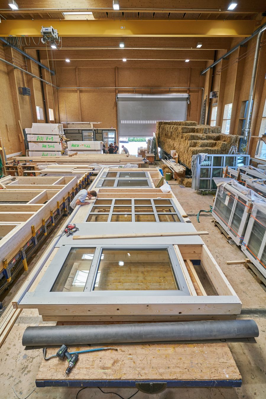  Das Bild zeigt eine große Werkhalle, in der Holzbau-Elemente gefertigt werden. Im Vordergrund liegen mehrere vorgefertigte Wandmodule mit eingebauten Fenstern auf Arbeitstischen. Die Rahmen bestehen aus Holz, und in einem Element ist eine Person zu sehen, die präzise Mess- oder Montagearbeiten durchführt. Auf dem Tisch liegen Werkzeuge wie ein Akkuschrauber, ein Bohrer und andere Bauutensilien. Im Hintergrund der Halle sind weitere Holzelemente gestapelt, ebenso wie große Ballen aus Stroh, die vermutlich als nachhaltiges Dämmmaterial verwendet werden. Rechts im Bild stehen fertig verpackte Bauteile, wahrscheinlich für den Transport oder die Montage vorbereitet. Die Halle selbst hat eine Holzkonstruktion mit hohen Decken, großen Fenstern und einer gelben Laufkrananlage an der Decke. Durch ein geöffnetes Rolltor am Ende fällt Tageslicht herein, und mehrere Personen arbeiten im hinteren Bereich. Insgesamt vermittelt das Bild den Eindruck einer modernen, nachhaltigen Holzbauproduktion mit Fokus auf ökologische Materialien und präzise Vorfertigung.