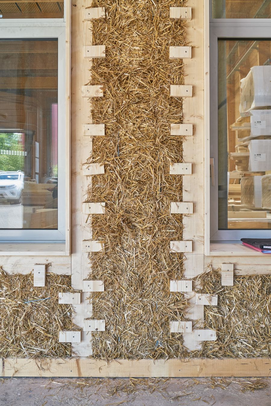 Das Bild zeigt eine Wandkonstruktion aus Holz und Stroh in einer Werkhalle, die sich offenbar im Bau oder in der Demonstrationsphase befindet. In der Mitte der Wand ist eine dichte Schicht aus gepresstem Stroh sichtbar, das als natürliches Dämmmaterial dient. Es ist zwischen hölzerne Rahmen eingefügt und wird von kleinen Holzklötzen fixiert, die regelmäßig angeordnet sind und das Stroh stabilisieren. Links und rechts befinden sich Fenster mit hellen Holzrahmen, durch die man sowohl ins Innere der Halle als auch nach draußen sehen kann. Im Inneren sind weitere Baumaterialien und Holzelemente zu erkennen. Der Aufbau zeigt anschaulich den ökologischen Konstruktionsansatz: eine Kombination aus Holz als tragendem Rahmenmaterial und Stroh als nachhaltige, umweltfreundliche Wärmedämmung. Die Szene vermittelt den Eindruck von innovativem, ressourcenschonendem Bauen im Holzbauhandwerk.