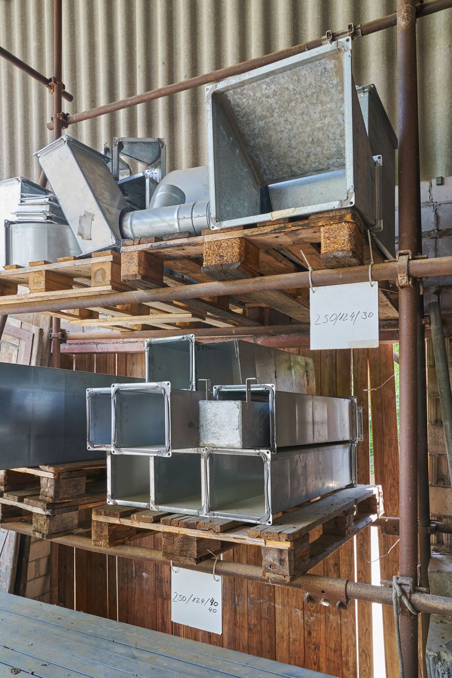 Das Bild zeigt ein Lagerregal mit verschiedenen Metallkanälen und Bauteilen, die auf Holzpaletten gestapelt sind. Die Teile bestehen aus verzinktem Stahlblech und haben unterschiedliche Formen und Größen – darunter rechteckige, quadratische und trichterförmige Elemente. Es handelt sich vermutlich um Komponenten für Lüftungs- oder Klimaanlagen, wie Luftkanäle und Verbindungselemente. Die Paletten sind in zwei Ebenen auf einem stabilen Gerüst aus Stahlrohren untergebracht. An den Ebenen hängen handbeschriftete Schilder mit Maßangaben, wie „250/124/40“ und „250/429/30“, wahrscheinlich zur Kennzeichnung der jeweiligen Bauteile. Der Hintergrund zeigt eine Holzwand und ein Wellblechdach, was auf ein Werkstatt- oder Lagerumfeld hinweist. Das Licht fällt natürlich von der Seite ein, was eine klare, strukturierte Arbeitsumgebung erkennen lässt.