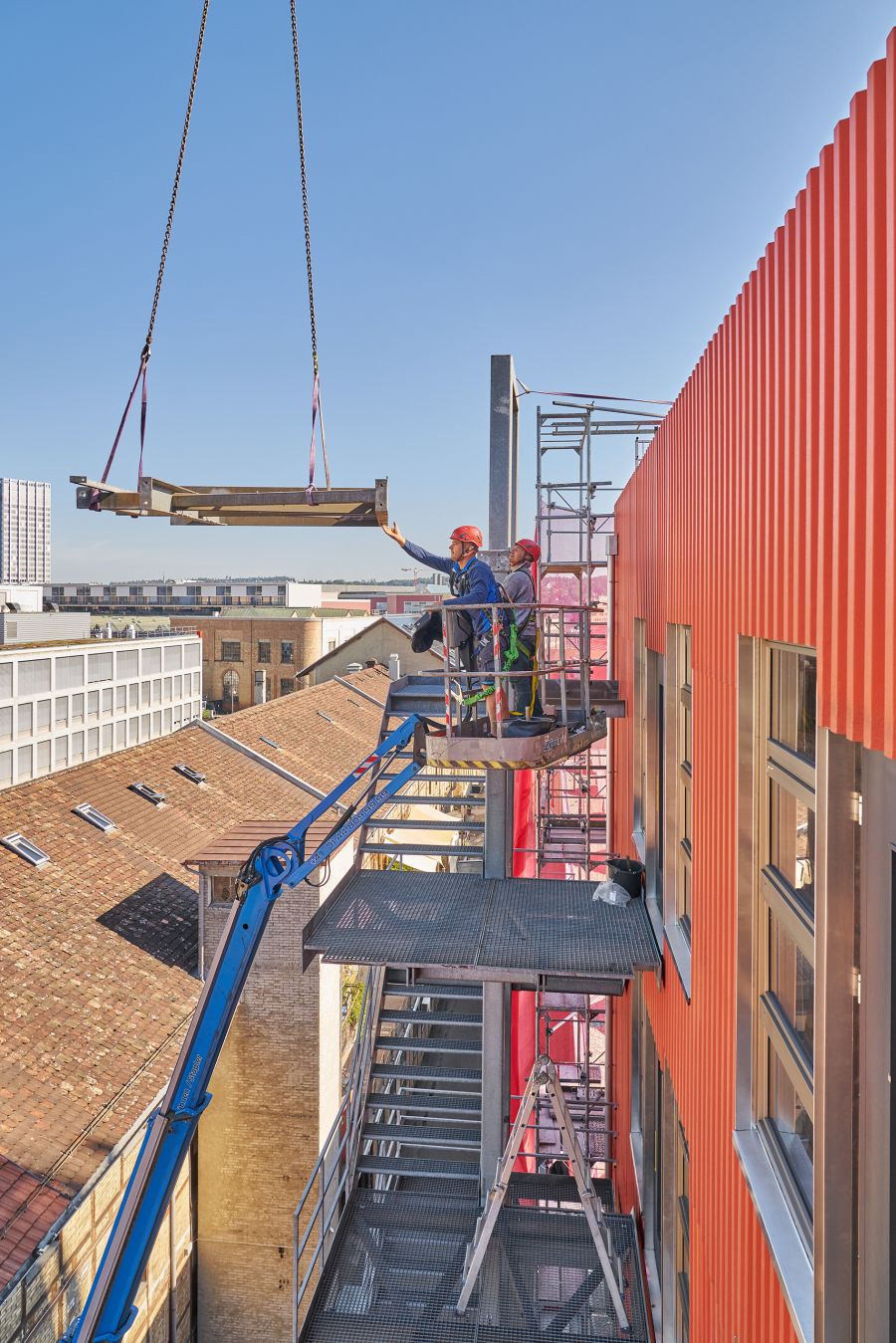 Das Bild zeigt eine Baustellenszene an der Fassade eines modernen Gebäudes mit orangefarbener Verkleidung. Zwei Bauarbeiter mit Sicherheitshelmen und Schutzgurten stehen auf einer Arbeitsplattform, die von einem blauen Hubarm getragen wird. Sie koordinieren das Anheben eines großen Stahlträgers, der mit Gurten und Ketten an einem Kran befestigt ist und gerade in Position gebracht wird. Einer der Arbeiter führt den Träger mit der Hand vorsichtig an seinen vorgesehenen Platz. Rechts im Bild ist die Fassade des Gebäudes mit vertikaler Wellblechstruktur und mehreren Fenstern zu sehen. Links erkennt man die Dächer älterer Nachbargebäude, während im Hintergrund ein hoher Turm und weitere moderne Bauten die Stadtsilhouette prägen. Der Himmel ist klar und blau, was auf einen sonnigen Tag hinweist. Die Szene vermittelt ein Bild von präziser Teamarbeit und moderner Baupraxis in luftiger Höhe.