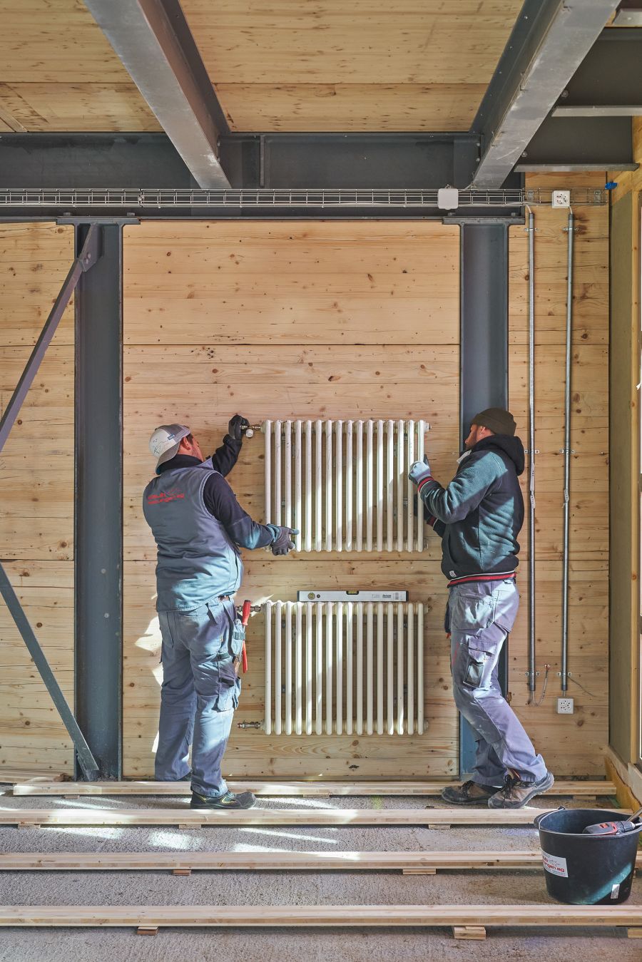 Das Bild zeigt zwei Handwerker, die in einem Innenraum einen Heizkörper an einer Wand montieren. Die Wand besteht aus hellem, unbehandeltem Holz, das zwischen dunklen Stahlträgern eingebaut ist – eine typische Kombination bei modernen Holz-Stahl-Konstruktionen. Die Arbeiter tragen Arbeitskleidung, Handschuhe und Mützen, und sie sind dabei, den oberen Heizkörper mit Werkzeugen an der Wand zu befestigen. Unter dem montierten Element ist bereits ein weiterer Heizkörper installiert, zwischen ihnen liegt eine Wasserwaage, um die exakte Ausrichtung sicherzustellen. Auf dem Boden befinden sich Holzlatten, vermutlich als Unterkonstruktion für einen späteren Bodenbelag. Rechts im Bild steht ein schwarzer Eimer mit Baumaterialien oder Werkzeugen. Die Umgebung ist hell und ordentlich, mit sichtbaren Leitungen und Kabeln entlang der Wand – ein typischer Anblick während der Ausbauphase eines Neubaus oder einer Sanierung.