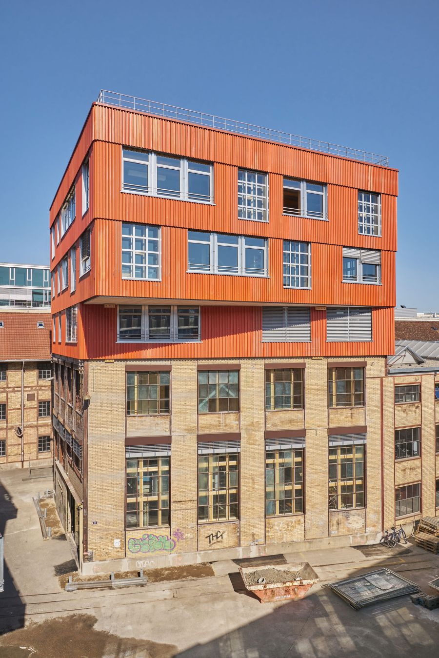 Das Bild zeigt ein mehrstöckiges Gebäude mit einer auffälligen architektonischen Kombination aus Alt- und Neubau. Der untere Teil des Gebäudes besteht aus gelb-beigem Backsteinmauerwerk mit großen, rechteckigen Industriefenstern. Diese Fassade erinnert an ein ehemaliges Fabrik- oder Lagerhaus. An der unteren Wand sind einige Graffiti zu erkennen. Der obere Teil des Gebäudes hebt sich deutlich ab: Er ist modern gestaltet und in einem kräftigen Orange gestrichen. Die Fassade besteht aus senkrechten Metallpaneelen und die Fensterrahmen sind silbergrau. Dieser neuere Abschnitt wirkt, als sei er auf das alte Gebäude aufgesetzt worden, was einen starken Kontrast zwischen historischer und zeitgenössischer Architektur erzeugt. Im Vordergrund ist ein Hof oder Innenhof mit Betonboden zu sehen, auf dem sich Baumaterialien, ein Container und ein Fahrrad befinden. Der Himmel ist klar und blau, was auf sonniges Wetter hinweist.