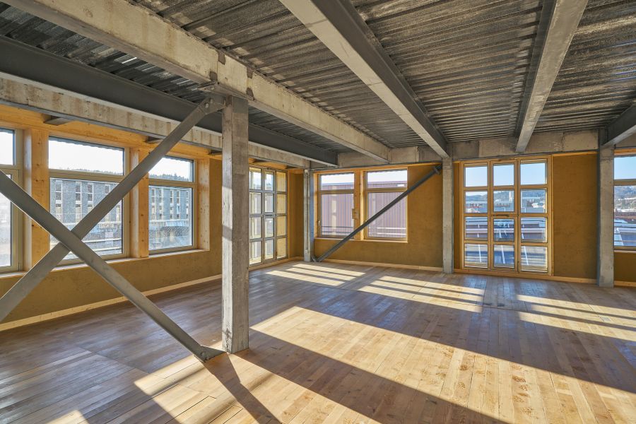Das Bild zeigt einen großen, offenen Raum mit industriellem Charakter. Die Wände bestehen aus einer Kombination von Beton, Stahl und Holzrahmen, die den Fenstern Struktur geben. Die Decke ist aus sichtbarem Metall mit Trägern und Verstrebungen, was dem Raum einen modernen, industriellen Look verleiht. Der Boden ist aus hellem, poliertem Holz gefertigt und reflektiert das Tageslicht, das durch die zahlreichen großen Fenster hereinfällt. Diese Fenster lassen viel natürliches Licht in den Raum und bieten Ausblicke auf umliegende Gebäude und eine leicht erhöhte Umgebung, möglicherweise ein städtisches Viertel oder ein Gewerbegebiet. Insgesamt wirkt der Raum hell, offen und roh – eine Mischung aus industrieller und moderner Architektur, die sich gut als Loft, Atelier, Büro oder Ausstellungsraum eignen würde.