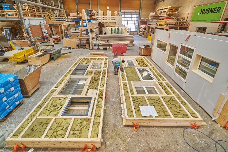 Das Bild zeigt das Innere einer großen Werkhalle, in der Holzelemente für ein Gebäude vorgefertigt werden. Die Halle ist hoch, gut beleuchtet und voller Maschinen, Holzstapel und Baumaterialien. Im Vordergrund liegen zwei große, flache Holzrahmenkonstruktionen nebeneinander auf dem Boden. Es handelt sich um vorgefertigte Wandmodule. Die Rahmen bestehen aus hellem Holz und sind bereits mit mehreren eingebauten Fenstern versehen. Zwischen den Holzbalken liegt grob zugeschnittene, grünlich-gelbe Dämmwolle, die in die offenen Fächer der Wandkonstruktion eingelegt wird. Zwischen den beiden Wandmodulen kniet ein Arbeiter. Er trägt Arbeitskleidung und einen Schutzhelm. Er arbeitet konzentriert daran, Dämmmaterial in einen der Holzrahmen einzufügen oder es zu glätten. Rechts neben ihm lehnt ein bereits fertig beplanktes Wandmodul senkrecht an einer Stütze. Es ist weiß verkleidet und mit eingebauten Fenstern versehen – unterschiedlich große Fenster, die sauber in einer glatten Fassade sitzen. Die Umgebung der Werkhalle wirkt geschäftig: Auf der linken Seite stehen mehrere Stapel von Holz und Pakete mit Baumaterialien, darunter blaue Säcke. Weiter hinten sieht man Arbeitsstationen, Tischsägen und Regale voller Holzlatten und Platten. Im Hintergrund befindet sich ein großes Tor, das vermutlich als Zufahrt für Material oder fertige Elemente dient. Rechts hängt ein großes grünes Schild mit dem Namen „HUSNER“, vermutlich der Name der Zimmerei oder des Herstellers. Der Gesamteindruck: Eine professionelle Holzbau-Werkstatt, in der Wandelemente in präziser Handarbeit und industriellem Umfeld vorgefertigt werden.