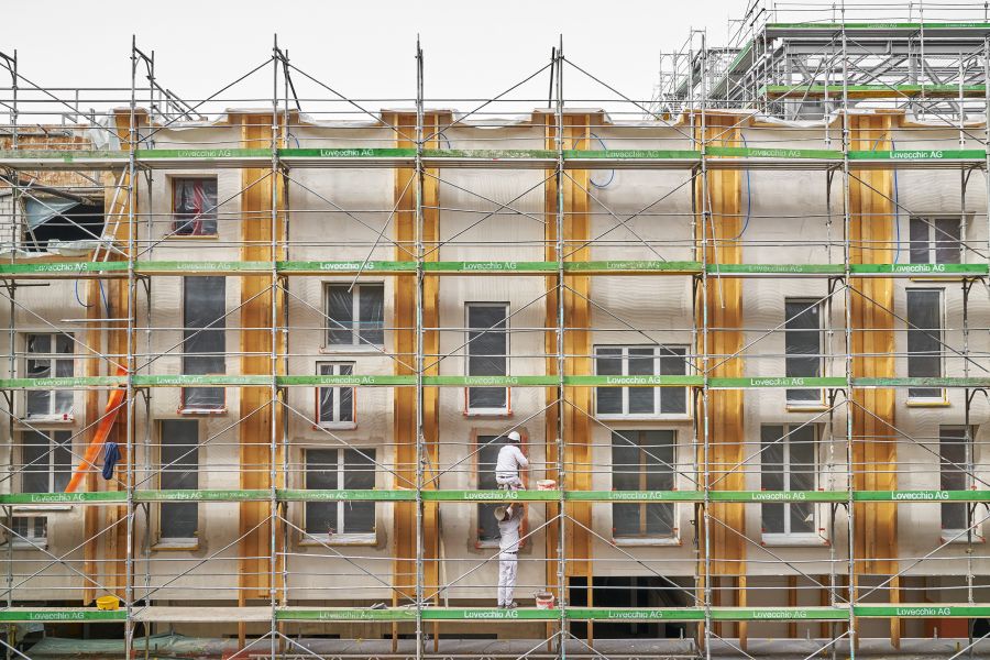 Das Bild zeigt die Fassade eines Wohnhauses während einer Sanierung, komplett umschlossen von einem Metallgerüst. Die hellen Wände sind mit auffälligen, vertikalen Streifen aus gelb-orangefarbenem Schutzgrund gestrichen. Viele Fenster sind mit Folien abgeklebt. Grüne Gerüstbretter tragen die Aufschrift „Lovecchio AG“. Auf einer unteren Ebene arbeitet ein Maler in weißer Kleidung und Helm mit einem Farbroller. Oben links führt ein orangefarbener Schuttrüssel vom Gebäude nach unten. Auf dem Gerüst sind Werkzeuge und Farbeimer verteilt. Die Szene vermittelt den Eindruck einer großflächigen, organisierten Fassadenrenovierung, bei der das Gerüst ein dichtes Raster vor der farblich strukturierten Wand bildet.