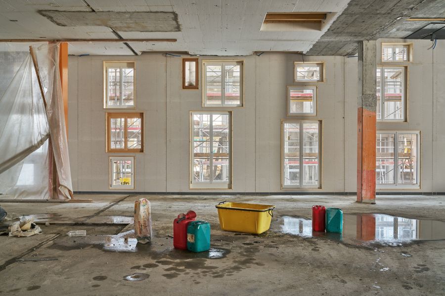 Das Bild zeigt den Innenraum eines Gebäudes, das sich mitten in einer Renovierung oder einem Umbau befindet. Der Raum ist groß, leer und wirkt unfertig. Der Boden besteht aus rohem Beton, auf dem sich mehrere Pfützen gebildet haben. Darin spiegeln sich Teile der Umgebung. Im Vordergrund stehen einige verstreute Gegenstände: Ein großer gelber Kunststoffbehälter, ein roter und ein türkisfarbener Kanister, ein aufgerissener Sack Baumaterial, vermutlich Zement oder Spachtelmasse. Alles wirkt provisorisch abgelegt, als hätten Bauarbeiter den Raum gerade verlassen. Die Wand im Hintergrund ist hell und durchzogen von vielen eingebauten Fenstern. Sie sind in verschiedenen Größen angeordnet – kleine quadratische Fenster, größere rechteckige und hohe bodentiefe Fenster. Die Rahmen sind aus hellem Holz. Durch die Scheiben sieht man außen ein Gerüst, was darauf hindeutet, dass auch die Fassade renoviert wird. Links hängt ein großes Stück transparente Baufolie locker herab, als eine Art Staubschutz. Rechts steht ein Betonpfeiler, im unteren Bereich orange gestrichen. Auch die Decke zeigt unverputzten Beton und mehrere offene Stellen, Kabel und Aussparungen. Der Gesamteindruck ist ein Baustellenraum im frühen Ausbauzustand – roh, nass vom Regen oder Bauwasser, mit verstreutem Material und vielen neuen Fenstern in einer sonst leeren Halle.
