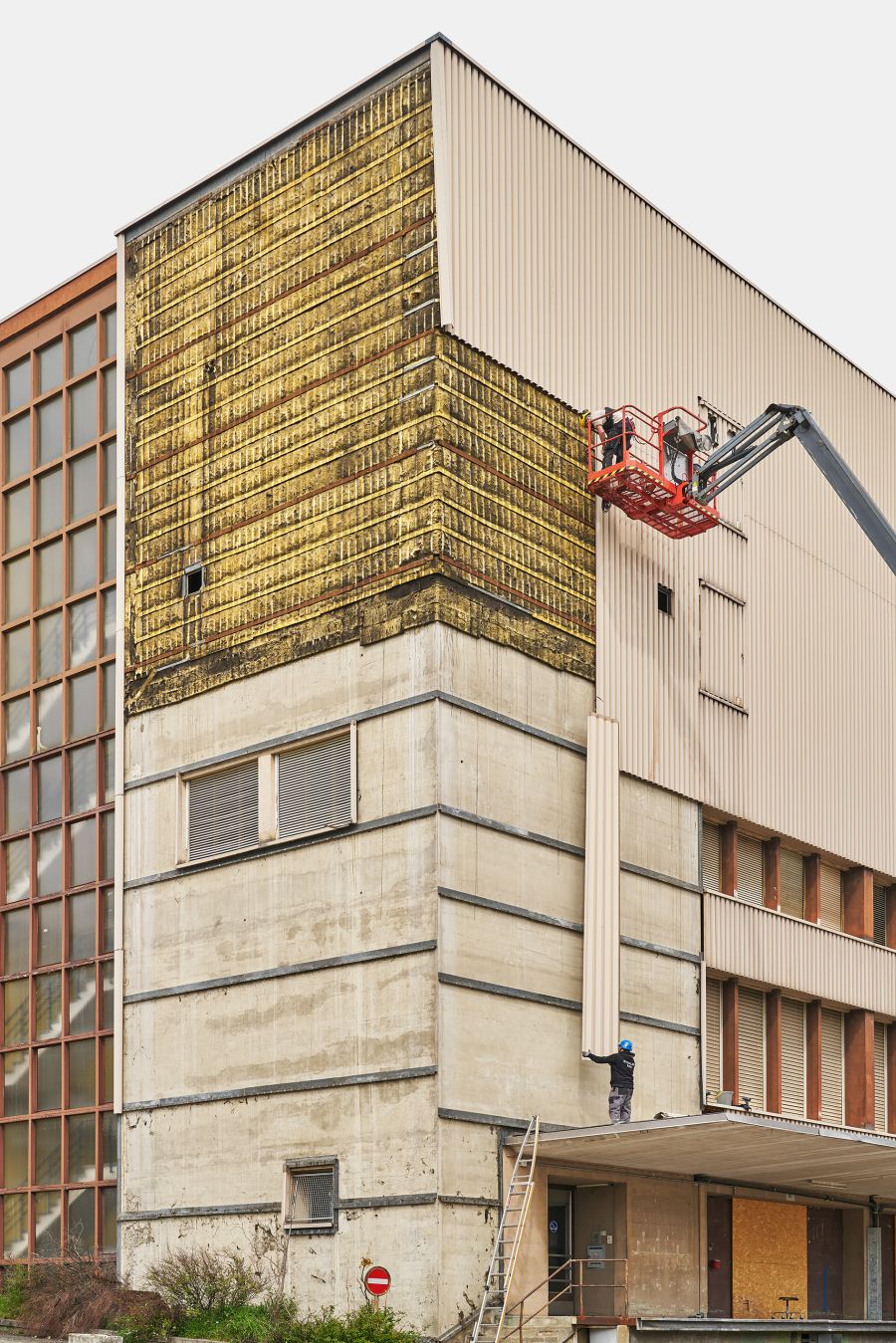 Das Bild zeigt ein Industrie- oder Gewerbegebäude, das sich im Umbau befindet. Im oberen Teil wird die alte, bestehende Fassadenverkleidung aus beigen, geriffelten Metallpaneelen abmontiert. Dadurch ist die darunter liegende gelbliche Isolierung sichtbar. Der untere Bereich der Fassadenverkleidung wurde bereits vollständig abmontiert und man sieht die Betonfassade. Rechts arbeitet ein Bauarbeiter in einer orangefarbenen Hebebühne an der Fassade. Ein weiterer Arbeiter steht darunter und hält ein Fassadenverkleidungselement. Der untere Gebäudeteil besteht aus rohem Beton mit horizontalen Schalungsspuren. Links erstreckt sich ein großes Fensterband mit rötlich-braunem Rahmen. Der Eingangsbereich wirkt verlassen und ist teilweise verbarrikadiert. Im Vordergrund sind Vegetation und ein „Einfahrt verboten“-Schild zu sehen. Das Bild vermittelt den Eindruck eines funktionalen Altbaus, der umfassend modernisiert wird.
