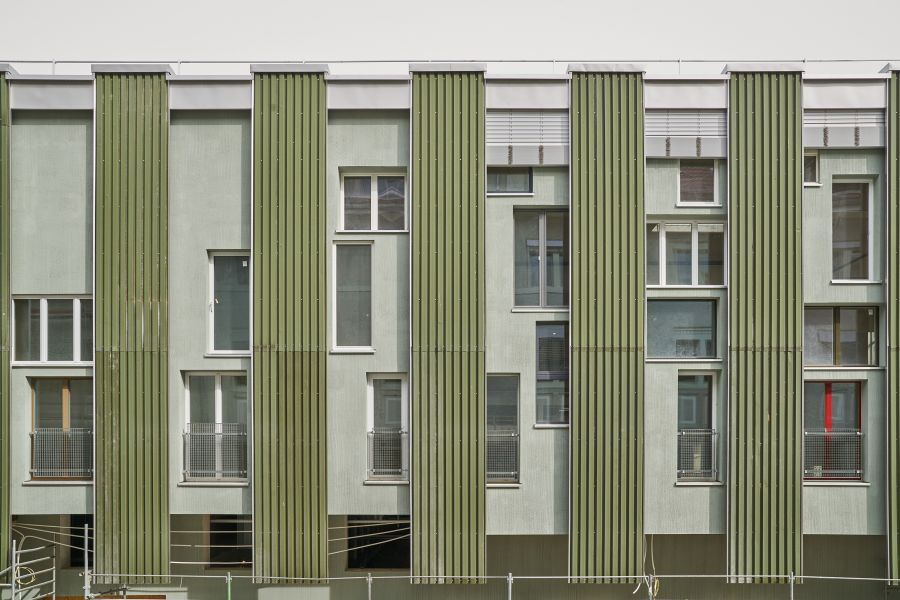 Das Bild zeigt die Fassade eines modernen Wohn- oder Bürogebäudes mit einem markanten, vertikal strukturierten Design. Die Außenwand ist in einem hellgrünen Ton gehalten und wird von fünf senkrechten grünen Lamellen unterbrochen, die optisch wie Paneele oder Fassadenverkleidungen wirken. Diese vertikalen Elemente schaffen ein rhythmisches, fast skulpturales Fassadenbild. Zwischen den Lamellen befinden sich Fenster unterschiedlicher Größen und Formate: Einige bodentief, andere kleiner oder als schmale Hochformate, zum Teil mit Gitterbrüstungen, die auf französische Balkone hinweisen könnten. Oben verläuft ein durchgehendes horizontales Bauteil, wahrscheinlich ein Dachabschluss oder eine technische Attika. Die gesamte Gestaltung kombiniert klare Linien, eine reduzierte Farbpalette und eine Mischung aus funktionalen und ästhetischen Elementen, was dem Gebäude ein zeitgenössisches, leicht industrielles Erscheinungsbild verleiht.