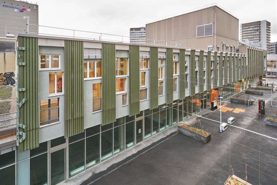 Diese Außenansicht zeigt ein langes, zweistöckiges Gebäude mit moderner Fassade. Das Obergeschoss ist in blassgrün gehalten und durch olivgrüne, senkrechte Paneel-Rippen rhythmisch gegliedert. Dazwischen liegen verschieden hohe Fenster, teils mit Brüstungen. Das Erdgeschoss besticht durch eine durchgehende, hohe Glasfront, die tiefe Einblicke ins Innere gewährt. Ein orangefarbener Eingang rechts setzt einen kräftigen Farbakzent. Vor dem Gebäude erstreckt sich ein weiter, leerer Asphaltplatz mit rechteckigen Pflanztitrogen, weißen Laternen und Bänken. Im Hintergrund ragen rechts ein Hochhaus und links ein Industriebau auf. Die Szenerie wirkt urban, sachlich und durch die klare Struktur der Fassade und des aufgeräumten Vorplatzes sehr funktional.