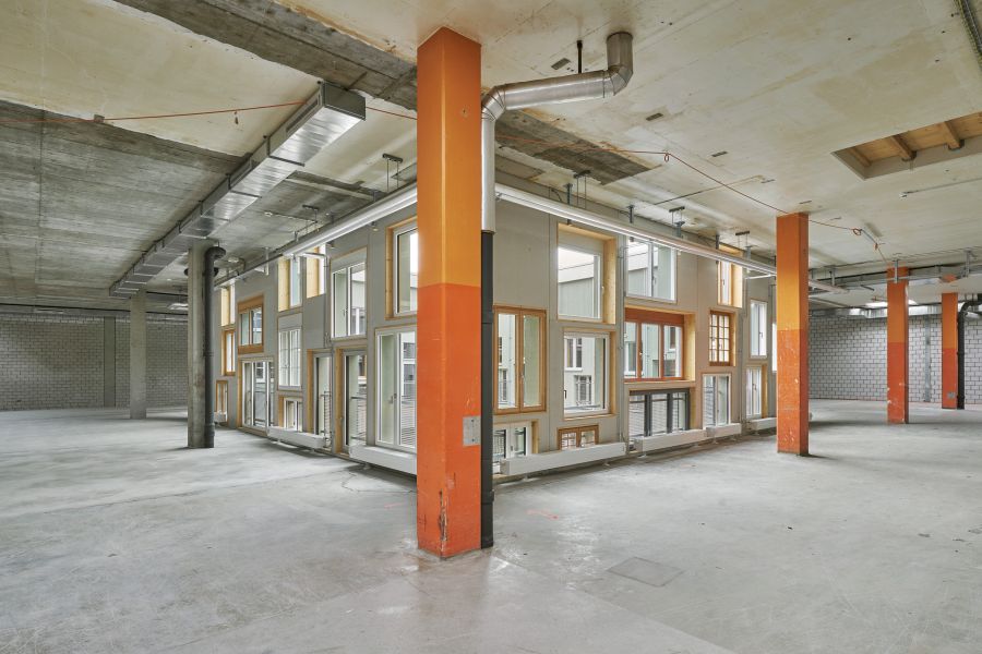 Das Bild zeigt einen großen, leeren Innenraum, der wie eine industrielle oder gewerbliche Fläche wirkt – möglicherweise ein ehemaliges Lager, eine Fabrikhalle oder ein Umbauprojekt. Die Decke und der Boden bestehen aus rohem Beton, und im Raum stehen mehrere kräftige, orange lackierte Stützpfeiler. Sichtbare Lüftungsrohre, Lüftungskanäle und Installationen verlaufen unter der Decke, was dem Raum einen technisch-industriellen Charakter verleiht. Auffällig ist eine Art „Haus im Haus“: Eine Konstruktion in der Mitte des Raumes, die aussieht wie eine Fassade mit vielen Fenstern, Türrahmen und verschiedenen Fensterformen – teils bodentief, teils kleiner. Diese Innenkonstruktion wirkt wie ein würfelförmiger Kern mit mehreren Stockwerksöffnungen, als hätte man ein Stück Wohnhaus innerhalb einer Fabriketage installiert oder nachgebaut. Der Raum ist sauber, aber leer, was darauf hindeutet, dass er für ein neues Nutzungskonzept umgebaut wurde.