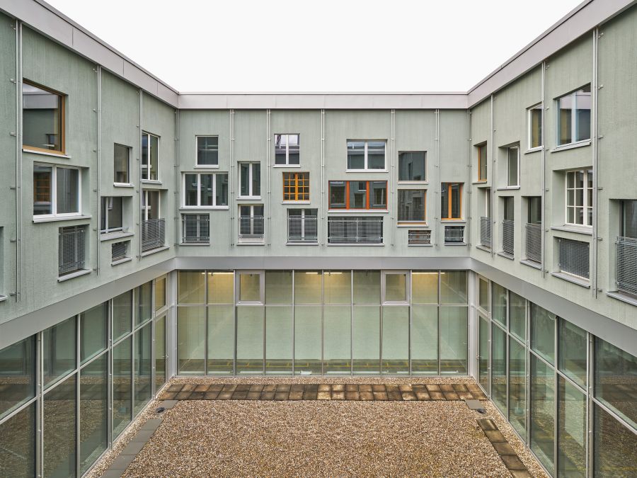 Das Bild zeigt einen rechteckigen Innenhof, der vollständig von einem modernen, mehrstöckigen Gebäude umschlossen ist. Der Betrachter blickt leicht erhöht in den Hof hinunter und auf die umliegenden Fassaden. Der Innenhof selbst ist ebenerdig und mit hellen Kieselsteinen bedeckt. In der Mitte verläuft ein schmaler, gerader Weg aus rechteckigen Betonplatten. Der Hof ist schlicht und ohne Pflanzen oder Möbel gestaltet. Das Gebäude rund um den Hof hat zwei Stockwerke. Das Erdgeschoss besteht fast vollständig aus großen Glasflächen: Breite Fensterfronten und Glastüren, die durch Aluminiumrahmen in rechteckige Felder unterteilt sind. Der Eindruck ist offen und transparent – man sieht in innenliegende Räume mit hellen Wänden und Deckenbeleuchtung. Das obere Stockwerk hat eine geschlossene Fassade in einem blassen Grün- oder Grauton. Überall sind Fenster eingebaut – sehr viele, in unterschiedlichen Formen und Größen: Hochrechteckige Fenster, breitformatige Fenster, manche mit weißen, manche mit Holzrahmen, einige mit kleinen Gitterbalkonen davor, die wie französische Balkone wirken. Die Fenster sind unregelmäßig, fast spielerisch angeordnet – nicht streng symmetrisch, sondern leicht versetzt, was dem Gebäude einen modernen, etwas lebendigen Charakter verleiht. Um den gesamten Innenhof herum zieht sich ein heller Balkon oder Gehweg auf Höhe des ersten Obergeschosses, unterbrochen von den Fenstergruppen. Der Gesamteindruck: ein ruhiger, sauberer, funktionaler Innenhof in einem zeitgenössischen Wohn- oder Bürogebäude, geprägt von vielen Fenstern, Glasflächen und einem schlichten, geordneten Innenraum.