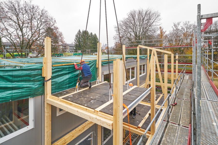 Auf dem Bild sieht man eine Baustellenszene im Freien. Im Vordergrund steht eine teilweise errichtete Holzkonstruktion, die an das bestehenden Gebäude angebaut ist. Die Struktur besteht aus hellen Holzstützen und einer Plattform aus dunklen Platten. Die Plattform ist noch an vier dicken Stahlketten gesichert, die vermutlich an einem Kran, der sich außerhalb des Bildes befindet, hängen. Rechts verläuft ein Metallgerüst mit rutschfesten Gitterböden. Eine grüne Plane ist um die Dachkante gespannt und schützt den Bereich darunter. In der Mitte kniet eine arbeitende Person auf der Plattform. Sie trägt Schutzkleidung: Einen Helm, eine Jacke und Handschuhe. Die Person befestigt oder überprüft die grüne Plane. Im Hintergrund stehen laublose Bäume, was auf Spätherbst oder Winter schließen lässt. Dahinter erkennt man einige Wohngebäude. Die gesamte Szene wirkt arbeitsintensiv und zeigt einen Zwischenstand im Holzbau.
