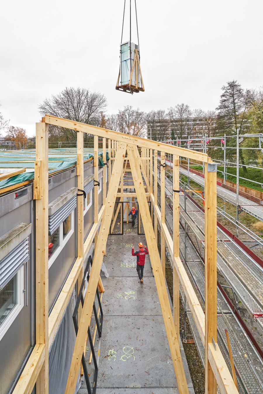 Das Bild zeigt eine Baustelle, betrachtet aus erhöhter Perspektive entlang eines schmalen Korridors innerhalb der errichteten Holzkonstruktion. Links steht ein graues Gebäude mit Fenstern, rechts verläuft ein langes Metallgerüst. Dazwischen befindet sich die neue Holzkonstruktion: Hohe, helle Holzpfosten und schräge Streben bilden ein Gerüst für zukünftige Wände und ein Dach. Oben in der Bildmitte hängt ein schweres Bauelement – ein gebündeltes Paket mit Fenstermodulen – an langen Ketten eines Krans. Es schwebt noch über der Baustelle und ist in einem Holzrahmen gesichert. Am Boden gehen zwei Bauarbeiter mit Schutzausrüstung. Einer blickt nach oben und hebt die Arme, vermutlich um das herabgelassene Element einzuweisen. Der Untergrund ist ein Betonboden mit gelben Markierungen. Im Hintergrund stehen herbstliche Bäume und einige Wohngebäude.
