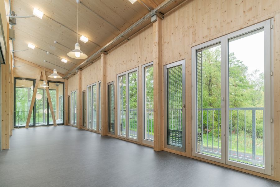 Man blickt, wie im oberen Bild ebenfalls dargestellt, in den Bereich der Klimazone. Das Bild zeigt den hellen, fertiggestellten Innenraum mit großer Fensterfront. Die Wände, Decke und tragenden Elemente bestehen aus hellem Holz, was dem Raum eine warme, natürliche Atmosphäre verleiht. Die Decke ist leicht geneigt und trägt mehrere runde, weiße Hängelampen. Entlang der rechten Seite und am hinteren Ende befinden sich große, bodentiefe Fenster und verglaste Türen. Einige sind mit außenliegenden Lamellen oder Gitterelementen versehen. Durch die Fenster sieht man viel Grün: Bäume, Sträucher und Wiesen. Der Boden ist dunkelgrau und wirkt glatt, vermutlich ein pflegeleichter Belag. Im hinteren Bereich erkennt man ein markantes, dreieckiges Holzstützelement, das wie ein großes „A“-Gestell aussieht. Der Raum ist leer und wirkt offen, freundlich und lichtdurchflutet.