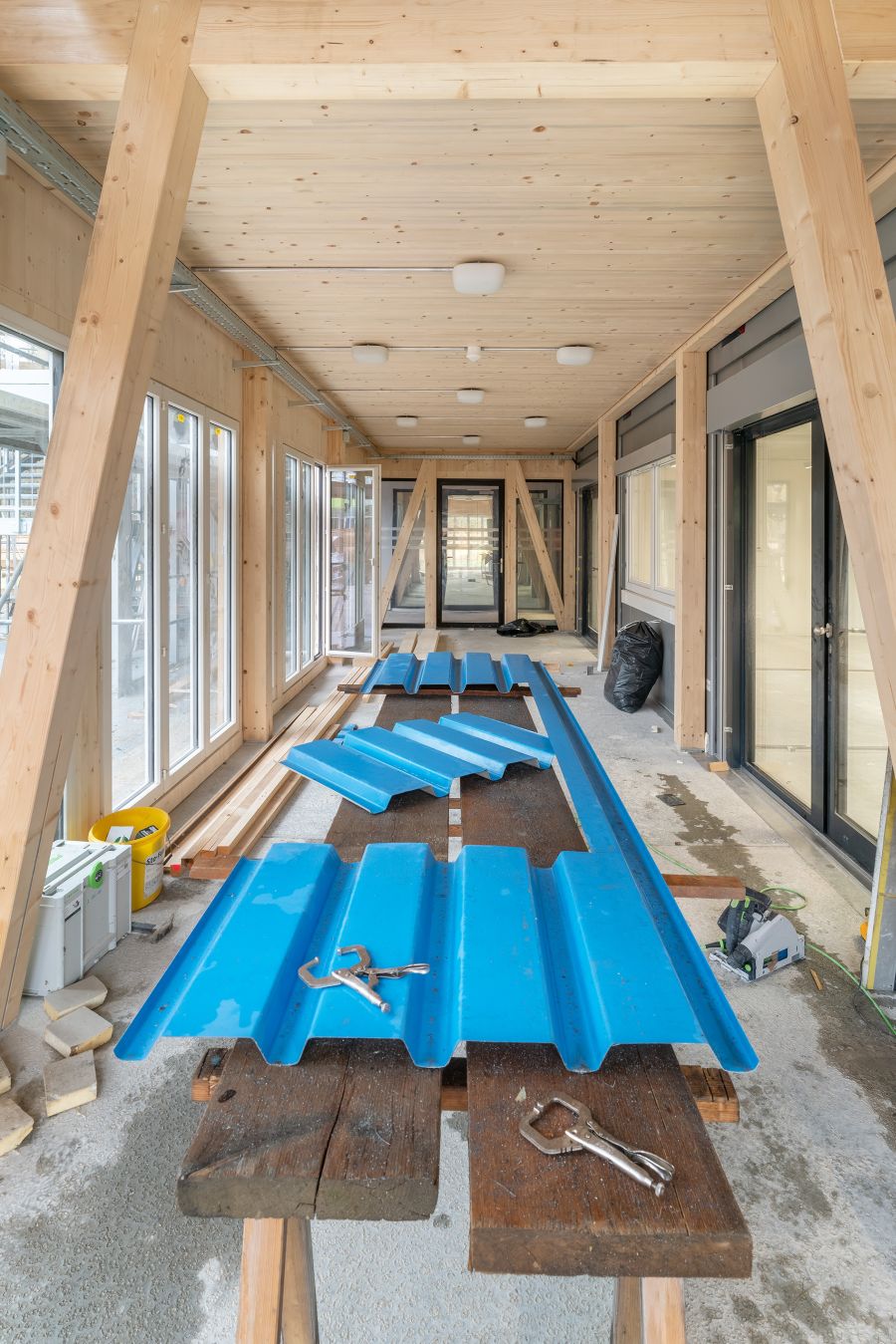 Man blickt in den Bereich der Klimazone. Das Bild zeigt einen langen, überdachten Gang innerhalb eines im Bau befindlichen Gebäudes. Die Konstruktion besteht überwiegend aus hellem Holz: Holzstützen, Holzwände und eine Holzdecke mit mehreren runden Lampen. Auf beiden Seiten des Gangs befinden sich große Fensterfronten und Glastüren. In der Mitte stehen zwei Holzböcke, auf denen mehrere lange, blau lackierte Metallbleche liegen. Sie sind leicht gewellt und offenbar Bauteile, die noch montiert werden sollen. Auf den Blechen liegen zwei Metallwerkzeuge, vermutlich Haken oder Greifer. Auf dem Boden liegen verschiedene Baumaterialien: Holzleisten, ein gelber Eimer, Werkzeugkoffer und ein schwarzer Müllsack. Der Boden wirkt roh und teilweise feucht, was auf laufende Arbeiten hindeutet. Am hinteren Ende des Gangs führt eine Glastür in einen weiteren Raum.