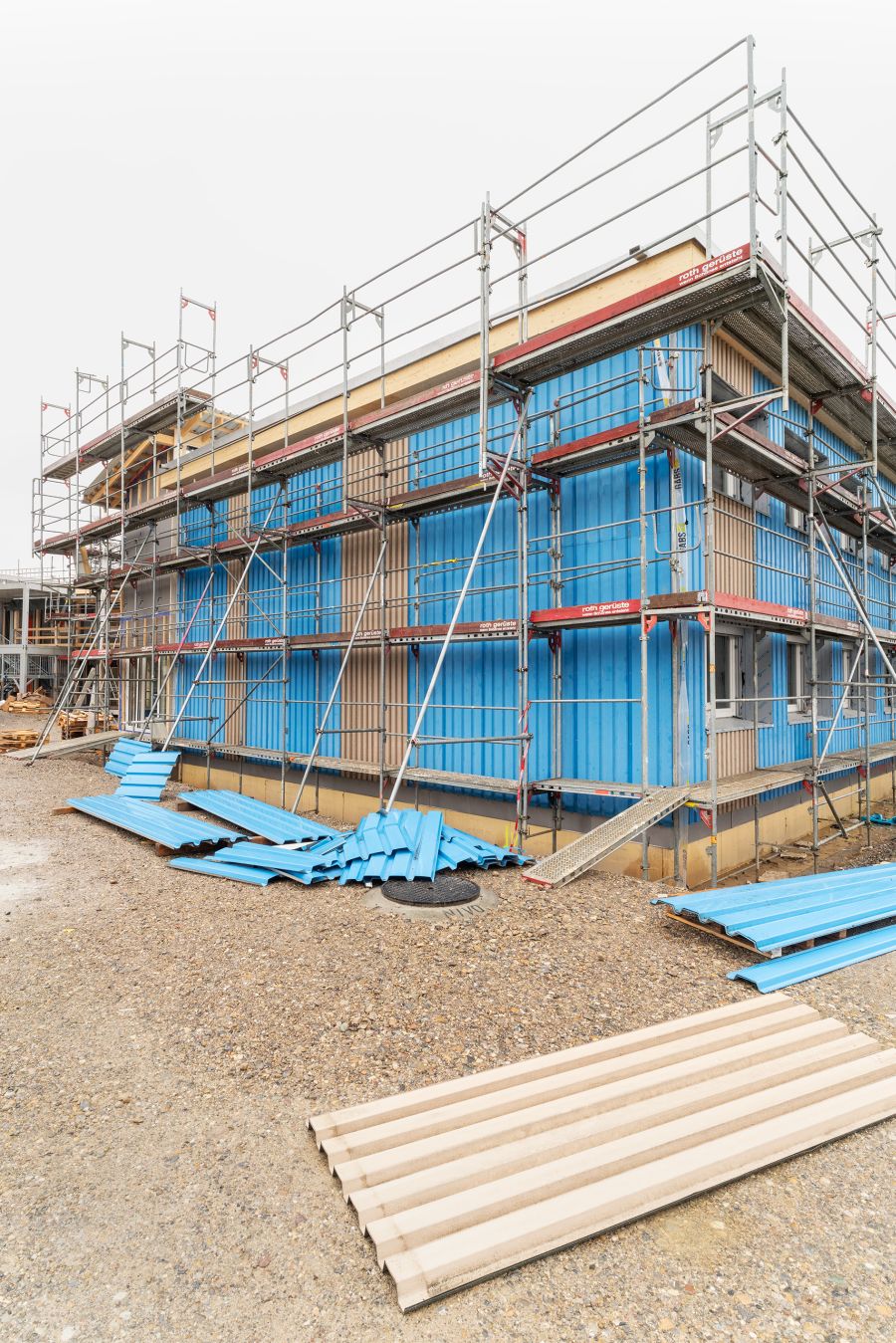 Das Bild zeigt ein Gebäude im Bauzustand, umgeben von einem hohen Metallgerüst. Die Fassade ist noch nicht fertiggestellt: Große, senkrecht montierte Platten in kräftigem Blau bedecken weite Teile der Außenwände. Zwischen einigen dieser blauen Elemente sind kleinere beige Paneele sichtbar. Vor dem Gebäude liegen auf dem kiesigen Boden mehrere Wellblechplatten – teils blau, teils beige –, die offenbar noch montiert werden sollen. Einige der blauen Platten sind aufgestapelt, andere liegen lose verteilt. Das Gerüst hat mehrere Ebenen mit Laufstegen und Schutzgeländern. Schräg gestellte Metallstützen stabilisieren die Struktur. Die Umgebung wirkt wie eine typische Baustelle: Offener Boden, Baumaterialien und im Hintergrund weiteres unfertiges Gebäudeteile. Alles deutet auf einen fortgeschrittenen Bauprozess hin, bei dem die Fassadenverkleidung montiert wird.