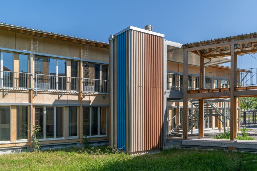 Das Bild zeigt einen Innenhofbereich eines modernen, zweistöckigen Holzbaus bei sonnigem Wetter. Die Fassade besteht überwiegend aus hellen Holzelementen und großen bodentiefen Fenstern oder Balkontüren. Entlang des oberen Stockwerks verläuft ein Metallgeländer, das als Absturzsicherung dient, unterstützt von Holzpfosten. Im Mittelpunkt steht ein markantes, vertikal geriffeltes Fassadenelement: Ein hoher, rechteckiger, fensterloser Schacht oder Technikraum, dessen Oberfläche aus senkrechten, farbigen Paneelen besteht. Diese Paneele sind in Blau, Beige und Rostbraun angeordnet und verleihen dem Gebäude einen lebendigen, gestreiften Akzent. Rechts befindet sich eine überdachte Passage aus Holz und Metall, die verschiedene Gebäudeteile miteinander verbindet. Darunter erkennt man eine Treppe, die in das obere Stockwerk führt. Unten im Innenhof wächst grünes Gras, das dem Bereich eine natürliche Note gibt. Der Himmel ist klar und blau, wodurch die Szene hell und freundlich wirkt.