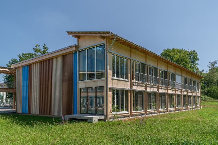 Das Bild zeigt ein modernes zweistöckiges Gebäude aus Holz, umgeben von einer grünen Wiese und Bäumen. Die Architektur wirkt leicht und offen. Die gesamte Front besteht aus großen, bodentiefen Fenstern oder Balkontüren über beide Stockwerke hinweg. Davor verläuft im oberen Geschoss ein Metallgeländer, das als Absturzsicherung dient. Links im Bild befindet sich eine markante Wandfläche aus vertikalen, farbigen Paneelen. Diese wechseln sich in Braun, Beige und Blau ab und verleihen dem Gebäude einen lebendigen, gestreiften Akzent. Die Dachlinie ist leicht geneigt, mit einem einfachen Blechdach. Unter dem Dach ragen Holzträger hervor, die die Konstruktion tragen. Rechts und im Hintergrund sieht man Bäume und Büsche, was auf eine ruhige, naturnahe Umgebung hindeutet. Das Gebäude wirkt fertiggestellt und modern, mit einem klaren Schwerpunkt auf Holzbau und viel Tageslicht.