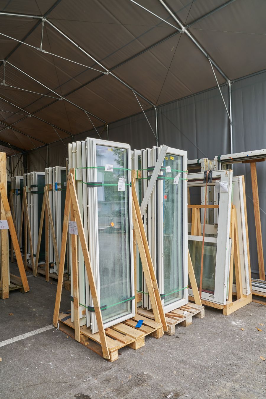 Das Bild zeigt eine überdachte Lagerfläche, in der zahlreiche gebrauchte oder vorbereitete Fensterflügel aus Glas und weißen Rahmen aufbewahrt werden. Die Fenster stehen auf Holzpaletten und sind jeweils in stabilen Holzgestellen fixiert, damit sie sicher und aufrecht gelagert werden können. Mehrere Gruppen solcher Fenster stehen dicht beieinander. Viele davon sind mit grünen oder schwarzen Gurten zusammengebunden. An manchen hängen Etiketten oder kleine Zettel, vermutlich mit Maßangaben oder Zuordnungen für den Einbau. Die Lagerfläche befindet sich unter einem großen Zeltdach aus dunklem Stoff, das von einem Gerüst aus Metallstangen gehalten wird. Der Boden ist asphaltiert, ähnlich einem Parkplatz. Insgesamt wirkt die Szene ordentlich organisiert und zeigt Materialien, die auf ihren Einsatz auf einer Baustelle warten.
