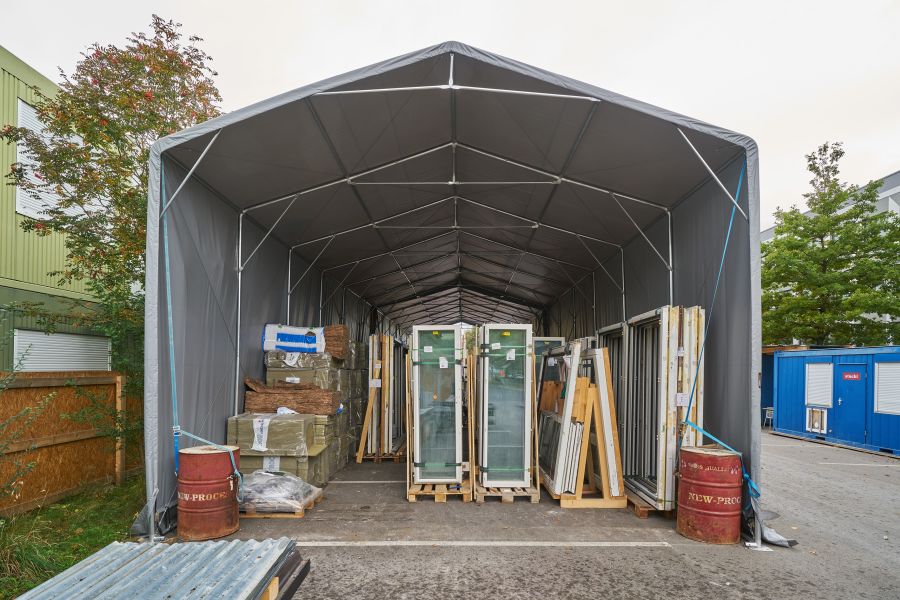 Das Bild zeigt ein großes, tunnelartiges Lagerzelt mit grauer Plane und einem Gerüst aus Metallstangen. Der Blick führt frontal in das Innere des Zelts. Im Inneren stehen verschiedene Baumaterialien ordentlich sortiert: In der Mitte mehrere hohe, stehende Fenster- oder Türrahmen mit Glas, jeweils auf Holzpaletten fixiert. Rechts daneben weitere Rahmen und Metallprofile. Links stapeln sich große Pakete mit Dämmmaterial und Bündel aus braunen Matten. Am Eingang des Zelts steht jeweils links und rechts ein altes, rotes Metallfass. Der Boden ist asphaltiert, wie auf einem Parkplatz. Außerhalb des Zelts sieht man links einen Holzzaun und einen Baum, rechts zwei blaue Container mit weißen Rollläden. Der Himmel ist hell, vermutlich bewölkt und die Szene wirkt wie ein geordneter Materiallagerplatz auf einer Baustelle. 