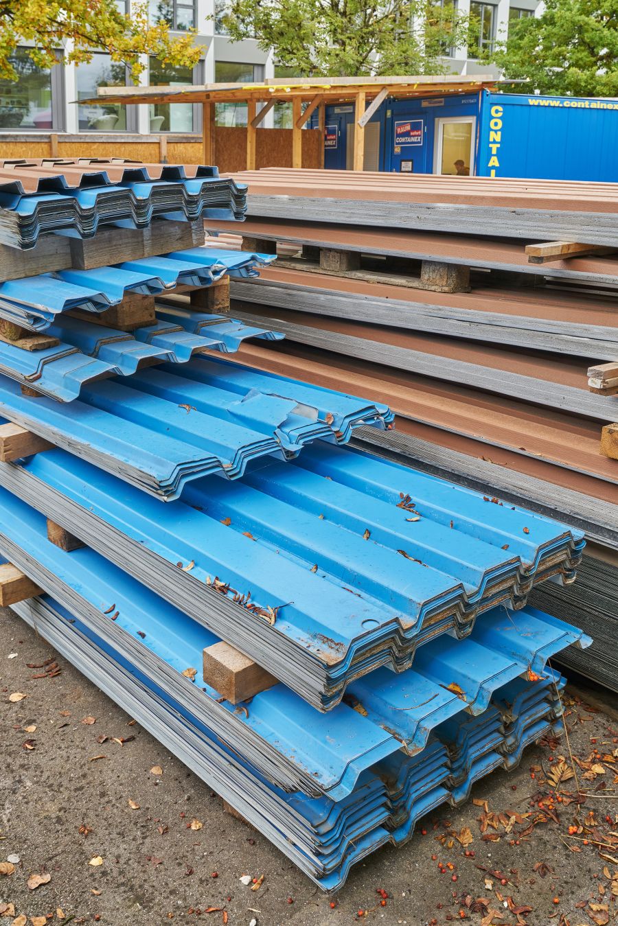 Das Foto zeigt einen Außenbereich eines Baustellen- oder Lagerplatzes. Im Vordergrund liegen mehrere gestapelte Metallplatten. Es handelt sich um große, gewellte oder trapezförmig profilierte Bleche, die typischerweise für Dächer oder Fassaden verwendet werden. Die oberen Bleche sind kräftig blau gefärbt, während weiter hinten auch graue und rotbraune Bleche zu sehen sind. Die Stapel liegen auf Holzpaletten und Holzklötzen, teils leicht unordentlich übereinandergelegt. Auf einigen Blechen liegen herabgefallene Blätter, was darauf hindeutet, dass Herbst ist. Der Boden besteht aus grobem Beton oder Asphalt und ist ebenfalls mit verstreuten Blättern und kleinen Früchten übersät. Im Hintergrund stehen einfache Holzkonstruktionen und ein blauer Container, der als Büro oder Lager dient. Auf dem Container sind Schilder zu sehen. Hinter all dem stehen mehrstöckige, moderne Gebäude mit großen Fenstern, teilweise durch Bäume verdeckt. Die Bäume haben gelb-grüne Blätter, was die herbstliche Stimmung verstärkt. Insgesamt vermittelt das Bild den Eindruck eines geordneten, aber rustikalen Baustellenlagers voller Baumaterialien.