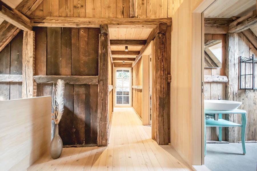 Das Foto zeigt den Innenbereich eines Hauses mit sehr rustikalem, warmem Flair. Es ist deutlich die Kombination aus altem, gebrauchten und neuem, hellen Holz zu erkennen. Man blickt in einen schmalen Flur aus hellem Holz. Die Wände, Decken und Balken bestehen überwiegend aus groben, dunklen und hellen Holzelementen. Man sieht die dunklen unbehandelt und teilweise rissigen Balken und Bretter, die vom ursprünglichen Gebäude erhalten geblieben sind. Der Dielenboden und einige Wände sind aus neuem, hellem Holz gefertigt. Links im Bild steht eine hohe, schmale Vase auf dem Boden, grau-glänzend, mit einigen dekorativen, hellen Gräsern darin. Rechts führt eine offene Tür in ein Badezimmer: Dort sieht man ein weißes rundes Waschbecken, das auf einem kleinen Tisch oder Gestell steht, das in einem pastell-türkisen Farbton gestrichen ist. Auch im Badezimmer bestehen die Wände aus den vom ursprünglichen Zustand des Hauses erhaltenen alten, groben Holzbalken. Am Ende des Flurs befindet sich eine Glastür oder ein großes Fenster mit Gittermuster, durch das natürliches Licht einfällt. Der gesamte Bereich wirkt freundlich, hell und gemütlich, mit einer Mischung aus modernem Minimalismus und traditionellem Holzhaus-Charakter.