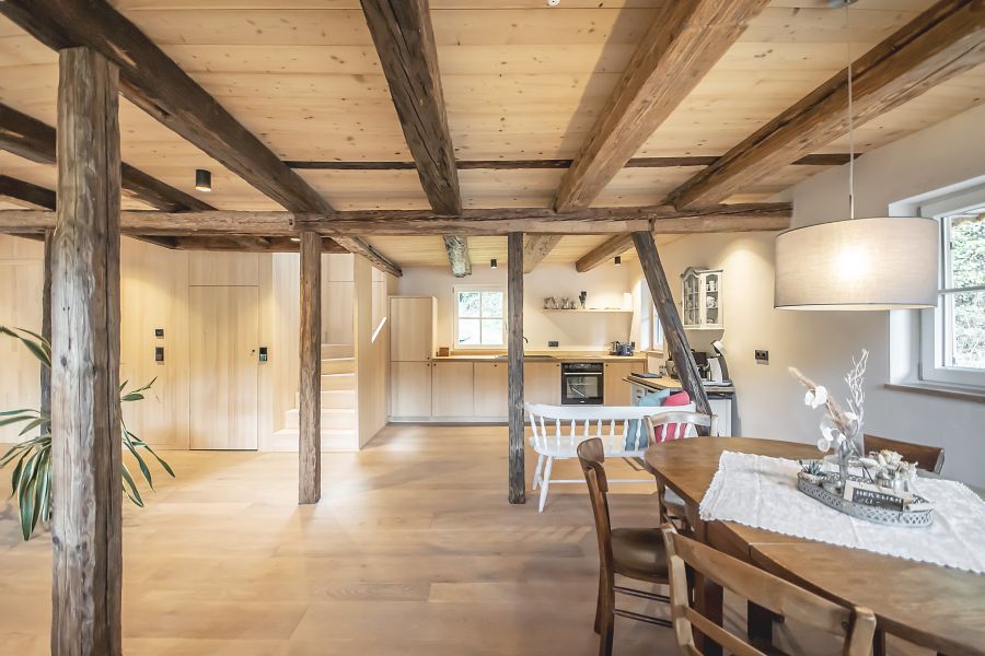 Das Foto zeigt einen offenen Wohnbereich in einem Haus, der im rustikalen Landhausstil gestaltet ist. Die Atmosphäre wirkt warm und gemütlich. Der gesamte Raum ist von hellen Holztönen geprägt: Fußboden, Wände und Decke bestehen aus neuem, hellem und glattem Holz. Quer über die Decke verlaufen die alten, ursprünglichen dunklen und massiven Holzbalken, die rustikal wirken. Einige ursprüngliche alte Balken stehen als Stützen frei im Raum, dicke, unregelmäßig geformte Stämme, die dem Raum einen traditionellen Charakter verleihen. Im Hintergrund befindet sich eine offene Küche. Die Küchenmöbel sind schlicht und ebenfalls aus hellem Holz. Es gibt einen Herd, eine Arbeitsfläche und darüber ein kleines Fenster, durch das Tageslicht einfällt. Im rechten Vordergrund steht ein ovaler Holztisch mit drei Stühlen. Auf dem Tisch liegt eine weiße, dekorative Tischdecke. Darauf steht ein Tablett mit einer dezenten Dekoration aus getrockneten Zweigen und kleinen Figuren. Darüber hängt eine große, runde, beige Stofflampe von der Decke herab. Links am Bildrand ist eine große grüne Zimmerpflanze zu erkennen. Im Raum stehen außerdem einige Möbelstücke im Landhausstil, darunter eine weiße Holzbank mit bunten Kissen. Der Gesamteindruck ist hell, freundlich und sehr natürlich, mit einer Mischung aus moderner Schlichtheit und traditionellem Holzbau.