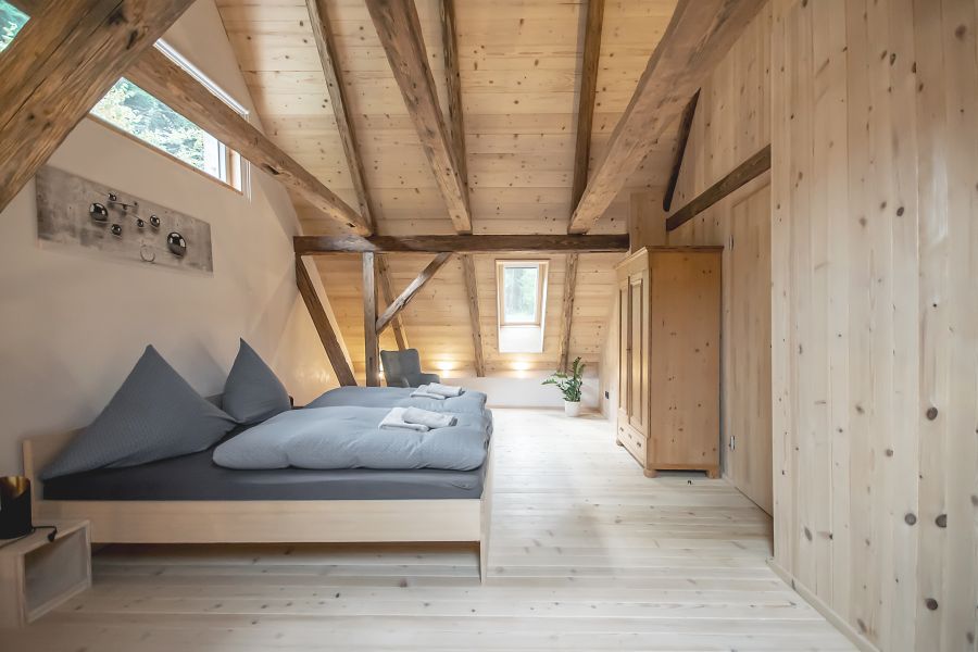 Das Bild zeigt ein helles, rustikales Schlafzimmer im Dachgeschoss, das hauptsächlich mit neuem, hellem Holz verkleidet ist. Links im Vordergrund dominiert ein großes, niedriges Doppelbett. Es hat einen schlichten, holzfarbenen Rahmen und ist mit einer graublauen Bettdecke und zwei großen, grauen Kissen bezogen. Auf dem Bett liegen ordentlich gefaltet zwei kleine, weiße Handtücher. Rechts vom Bett befindet sich eine freie Fläche aus hellem Dielenboden aus neuem Holz, gefolgt von einem hohen, zweitürigen Holzkleiderschrank in einer helleren Holzart. Im Hintergrund sieht man eine kleine Leseecke mit einem grauen Sessel. Die Wände sind teilweise mit glatten, neuen hellen Holzpaneelen und teilweise mit einer hellen, cremefarbenen Farbe versehen. Die Decke ist offen und zeigt die ursprünglichen alten, rustikalen und dicken Dachbalken aus dunklerem und unbehandeltem Holz. Diese alten Balken kontrastieren stark mit dem ansonsten sehr hellen, neuen Holz der Deckenverkleidung und des Fußbodens. Oben links in der Schräge befindet sich ein langes, schmales Fensterband. Ein kleineres, rechteckiges Dachfenster ist in der Mitte des Raumes, das einen Blick auf den Himmel oder eine andere Lichtquelle freigibt. Mehrere kleine, indirekte Strahler sind in der Nähe des Dachfensters installiert und beleuchten den Boden und die Wand. An der cremefarbenen Wand hinter dem Kopfteil des Bettes hängt ein abstraktes Kunstwerk mit silbernen, kreisförmigen Elementen auf einem hellen Hintergrund. Der Gesamteindruck ist hell, luftig und gemütlich mit einem starken Fokus auf natürliche Materialien und eine einfache, moderne Ästhetik.