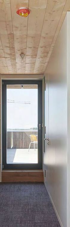 Der Raum ist ein schmaler, heller Flur mit grauem Teppichboden. Die Decke ist mit warmen, horizontalen Holzbohlen verkleidet. Ein kleines rundes Objekt mit rotem Rand, eventuell ein Rauchmelder, ist dort befestigt. Die rechte Wand ist eine glatte, helle Plattenwand mit einer modernen, bündig eingelassenen Tür mit silbernem Griff. Am Ende des Flurs schließt eine große, dunkle Glastür den Raum ab und führt zu einer Außenterrasse. Draußen ist helles Tageslicht durch eine blickdichte Holzbrüstung und Teile eines gelben Außenstuhls erkennbar. Die Atmosphäre wirkt klar, modern und kombiniert natürliche mit glatten, technischen Materialien.