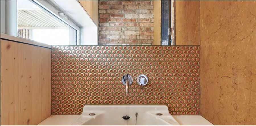 Diese Nahaufnahme zeigt ein Badezimmer mit kreativem Upcycling-Design. Blickfang ist die Wand über dem großen, weißen Waschbecken: Sie ist als Mosaik aus hunderten gold-beigen kronkorkenähnlichen Deckeln mit roten Akzenten gestaltet. Mittig ist eine moderne Einhandarmatur montiert. Links schließt eine helle Holzwand mit darüberliegendem Fenster an. Rechts besteht die Wand aus strukturiertem, naturfarbenem Lehm- oder Kalkputz. Über dem Mosaik ist ein Spiegel eingelassen, der eine gegenüberliegende Backsteinwand reflektiert. Das Design verbindet moderne Armaturen mit rustikalen Materialien und nachhaltiger Kreativität zu einem warmen, individuellen Raumerlebnis.