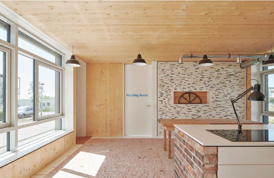 Dieser moderne Raum, möglicherweise ein Besprechungsraum oder eine offene Küche/Essbereich, kombiniert industrielles Design mit warmem Holz. Die Decke ist holzgetäfelt, der Boden hell gesprenkelt. Links lässt eine große Fensterfront viel Tageslicht herein; darunter verläuft eine tiefe Holzbank. Die Rückwand zeigt einen Mix aus Holzpaneelen, einer weißen Tür mit der Aufschrift „Meeting Room“ sowie Ziegel- und Betonelementen. In einer Nische ist ein hölzernes Wagenrad als Deko eingelassen. Im rechten Bereich stehen ein langer Holztisch und eine Kücheninsel mit weißer Platte und Ziegelsockel, auf der eine große schwarze Lampe steht. Von der Decke hängen industrielle Pendelleuchten; graue Rohre verlaufen sichtbar an der Wand. Das Ambiente wirkt offen und funktional durch die Verbindung von technischer Ausstattung und rustikalen Materialien.