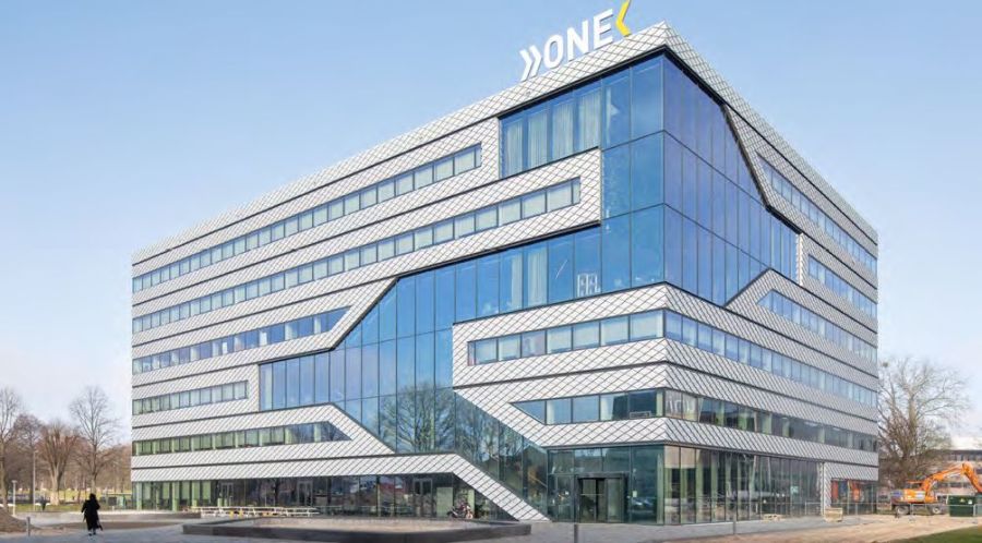 Das Bild zeigt das Matrix ONE Gebäude in Amsterdam, das sich durch seine markante, nachhaltige Architektur auszeichnet. Die Fassade ist durch eine Kombination aus großflächigen Glasfronten und schindelartigen, rautenförmigen Aluminiumelementen in weiß und hellgrau gekennzeichnet. Diese Elemente sind über die gesamte Gebäudehöhe in horizontalen Bändern angeordnet. Die horizontalen Bänder verlaufen in zickzackförmigen Linien diagonal über das Gebäude. Der Name 'ONE' ist in großen, weißen Buchstaben auf dem Dach des Gebäudes angebracht. Das Gebäude steht im Amsterdamer Science Park in einer Umgebung mit Bäumen, Gehwegen und einigen umliegenden Grünflächen. Im Vordergrund rechts ist ein kleiner Bagger auf einer Baustelle zu sehen. Der Himmel ist hellblau mit einigen weißen Wolken.